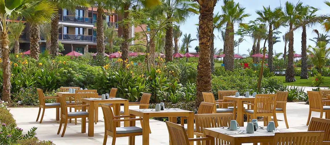Außenbereich des Hilton Cabo Verde Sal Resorts mit hölzernen Tischen und Stühlen unter roten Sonnenschirmen.