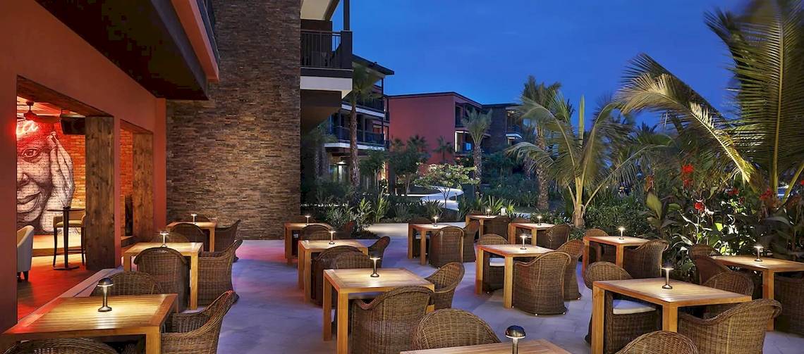 Außenbereich des Hilton Cabo Verde Sal Resorts bei Nacht mit beleuchteten Tischen und Stühlen, umgeben von Pflanzen und Gebäuden.