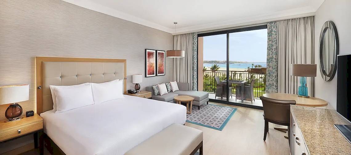 Innenansicht einer Suite im Hilton Cabo Verde Sal Resort mit großem Bett, Sofa und Balkon mit Meerblick.