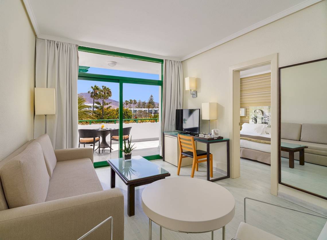 Innenansicht einer Suite im H10 Lanzarote Princess Hotel mit Wohnzimmer, Esstisch, Fernseher und Schlafzimmer.