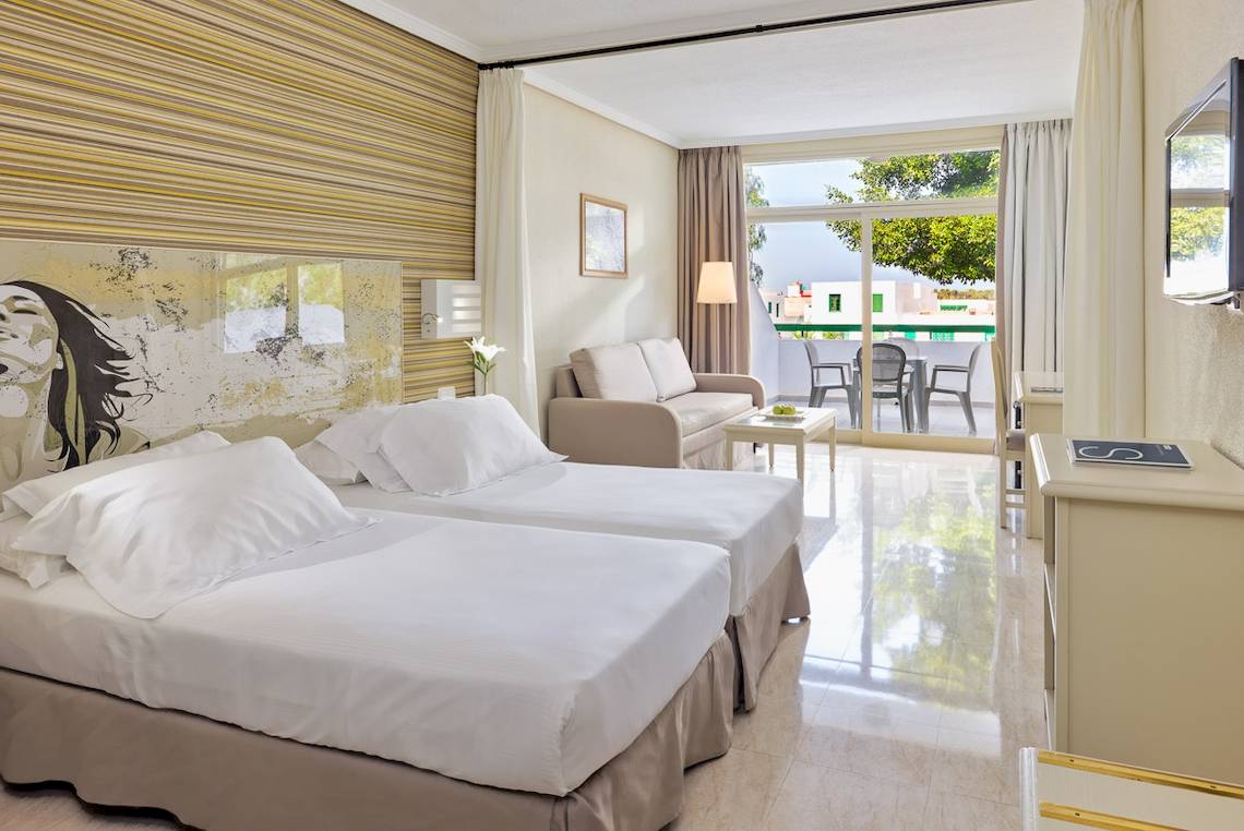 Innenansicht eines Doppelzimmers im H10 Lanzarote Princess Hotel mit zwei Betten, einem Sofa und einem Balkon.