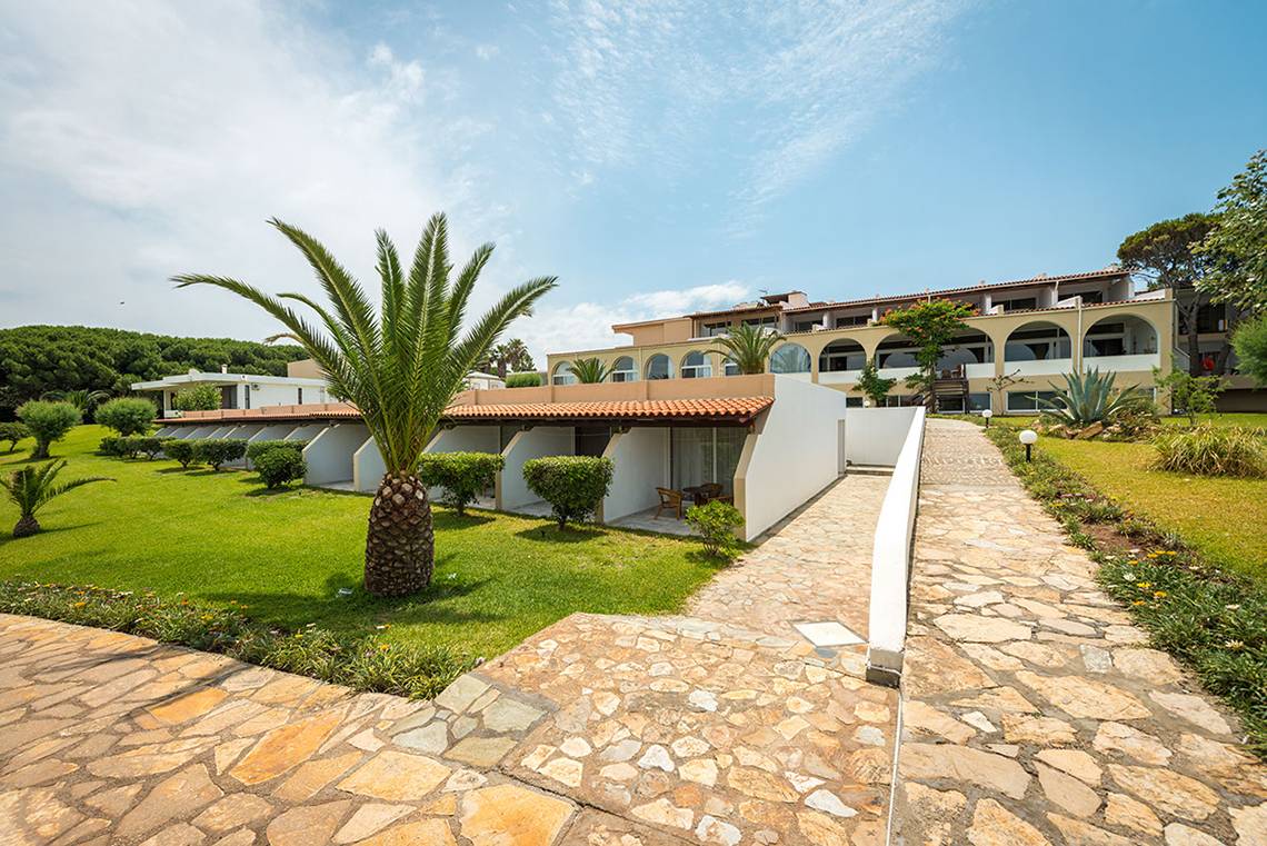 Außenbereich des Pavlina Beach Hotels mit gepflastertem Weg, Palme und Gebäude im Hintergrund.