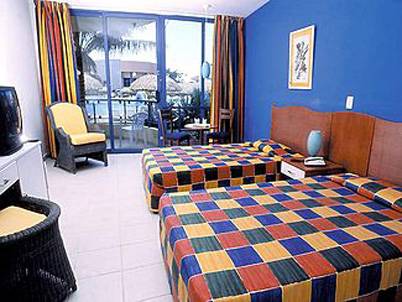 Doppelzimmer im Muthu Playa Varadero Hotel mit zwei farbenfrohen Betten, gelbem Stuhl und Fernseher.