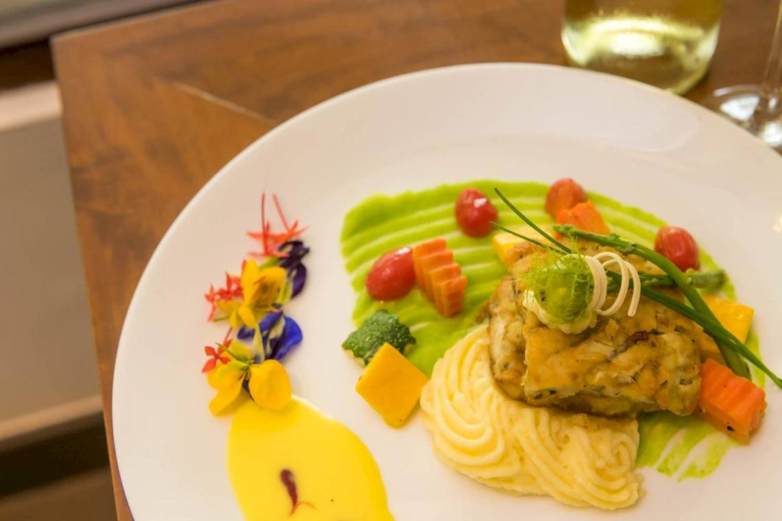 Ein Gericht auf einem weißen Teller, bestehend aus Fisch, Kartoffelpüree und Gemüse, mit einer grünen Sauce und dekorativen Blumen.