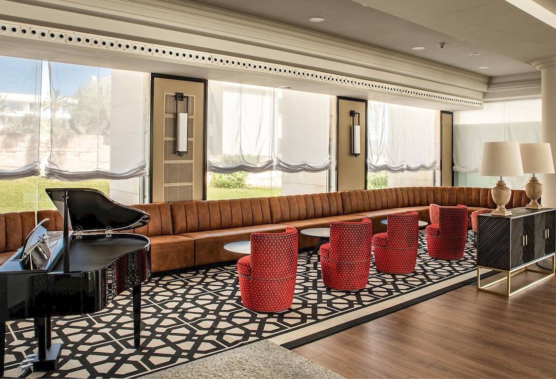 Hotellobby des Sarena de Muro Resorts auf Mallorca mit Klavier, roten Stühlen und großem Fenster.
