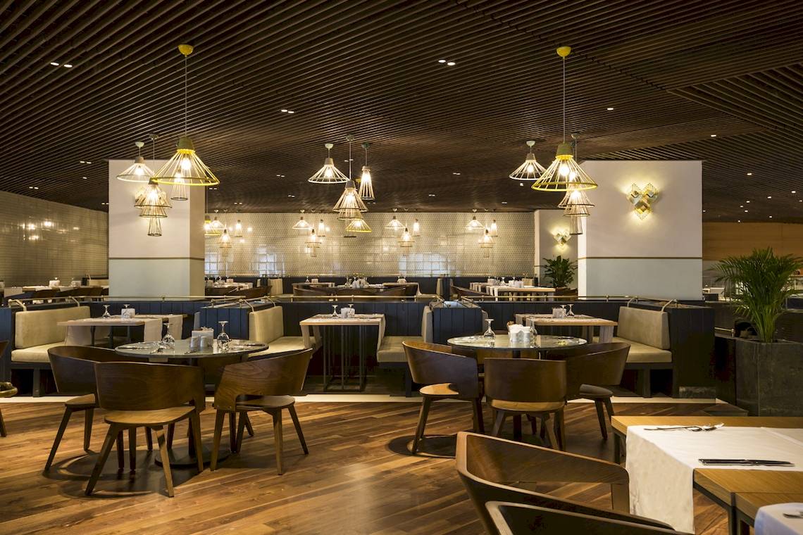 Innenbereich des Voyage Sorgun Hotels mit runden Tischen und Stühlen, Holzboden und hängenden Lampen.