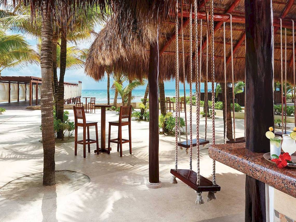 Strandbar des El Dorado Royale a Spa Resort by Karisma mit Tischen, Stühlen und einer Schaukel am Strand.