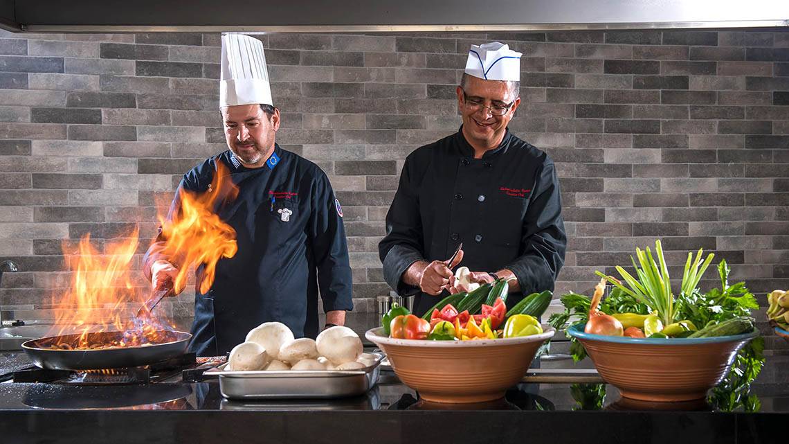 Zwei Küchenchefs des Alexander House stehen in der Küche und bereiten Essen zu, mit einem Koch, der Feuer benutzt, und Gemüse und Zutaten auf dem Tisch.