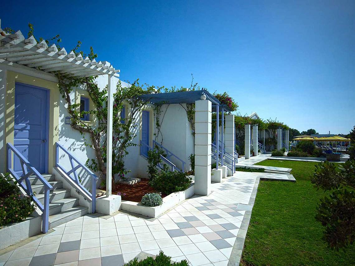 Außenbereich des Serita Beach Hotels mit Reihen von weißen Gebäuden, blauen Türen und Treppen, überdachten Gehwegen und grünem Rasen.