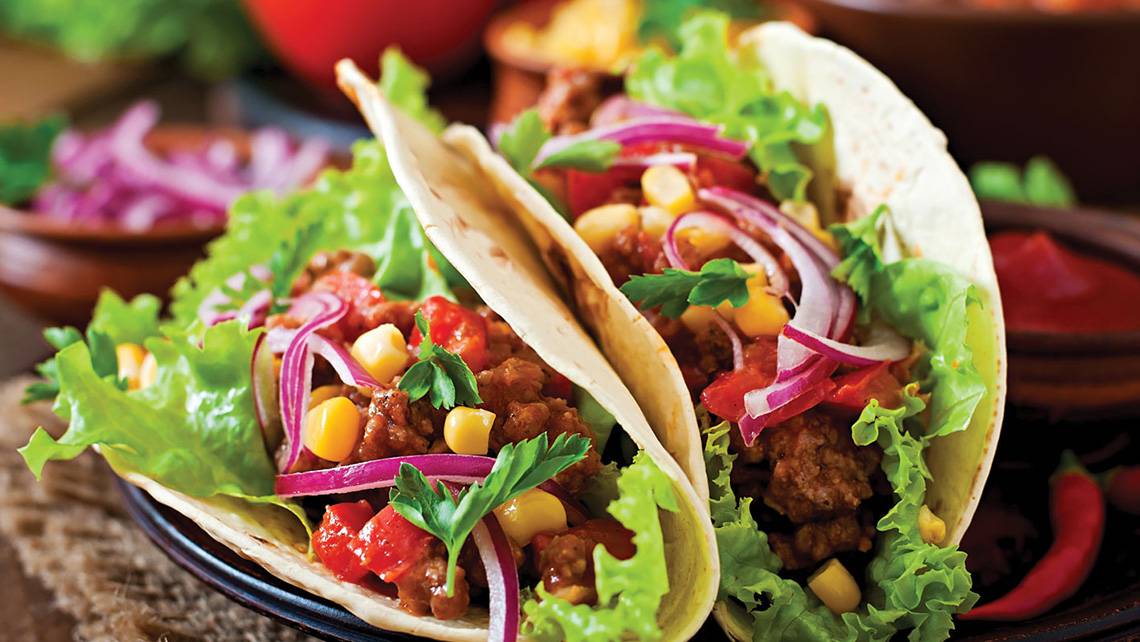Zwei Tacos mit Hackfleisch, Salat, Tomaten, Mais und roten Zwiebeln auf einem schwarzen Teller.