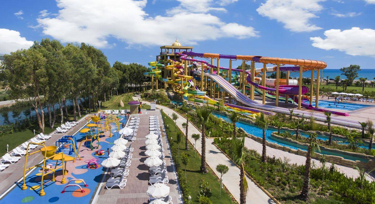 Wasserpark mit bunten Rutschen und Liegen im Delphin BE Grand Resort, umgeben von Grün und Himmel.