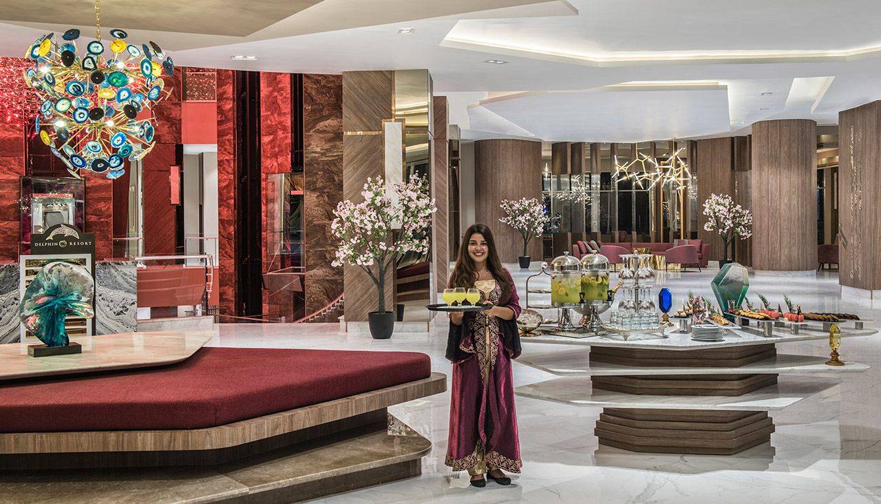 Elegante Hotellobby des Delphin BE Grand Resort mit einer lächelnden Kellnerin und einer farbenfrohen Deckenleuchte.