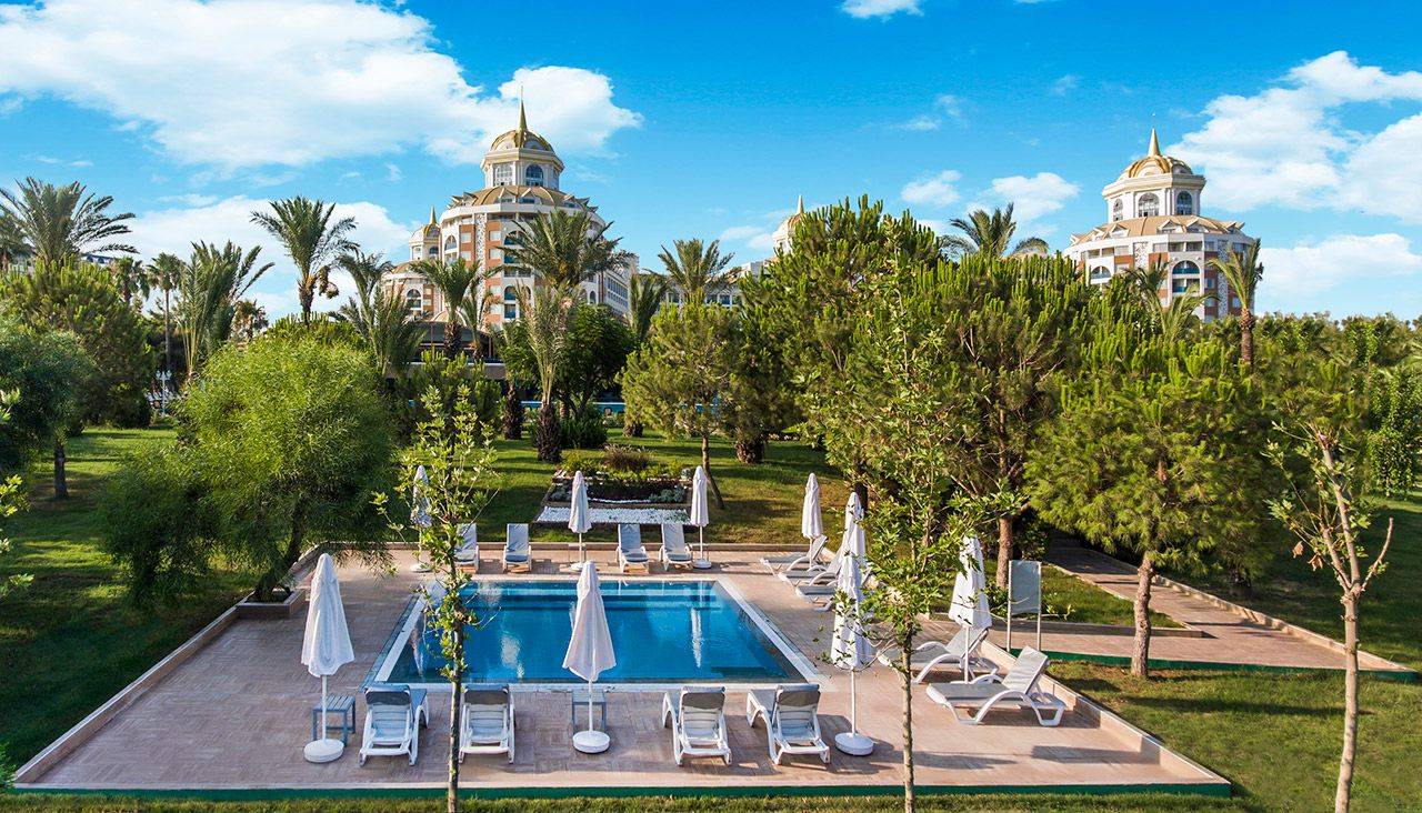 Ansicht des Delphin BE Grand Resort mit Pool und Palmen unter blauem Himmel. Entspannte Atmosphäre im Garten.