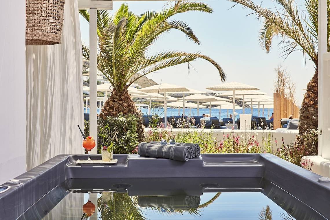 Poolbereich des Mykonos Ammos Hotels mit Blick auf die Palmen, Sonnenschirme und das Meer.