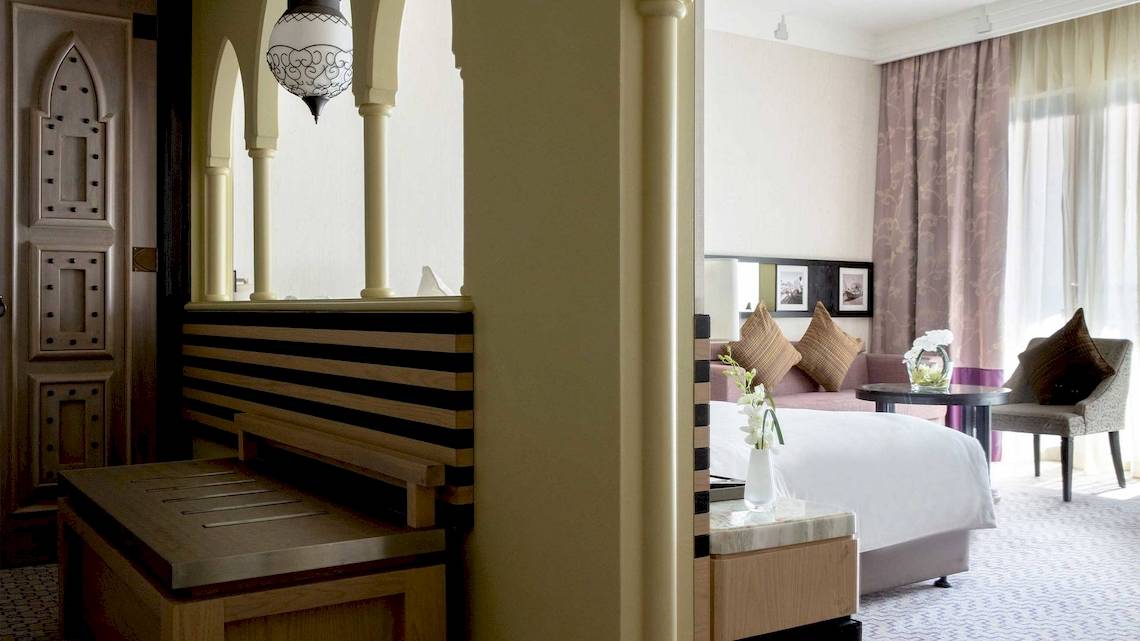 Innenansicht des Jumeirah Mina A'Salam Hotelzimmers mit Bett, Sitzbereich und Fenstern.