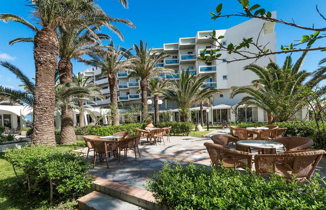 Außenbereich des Apollo Beach Hotels mit Palmen, Tischen und Stühlen vor einem modernen Gebäude.