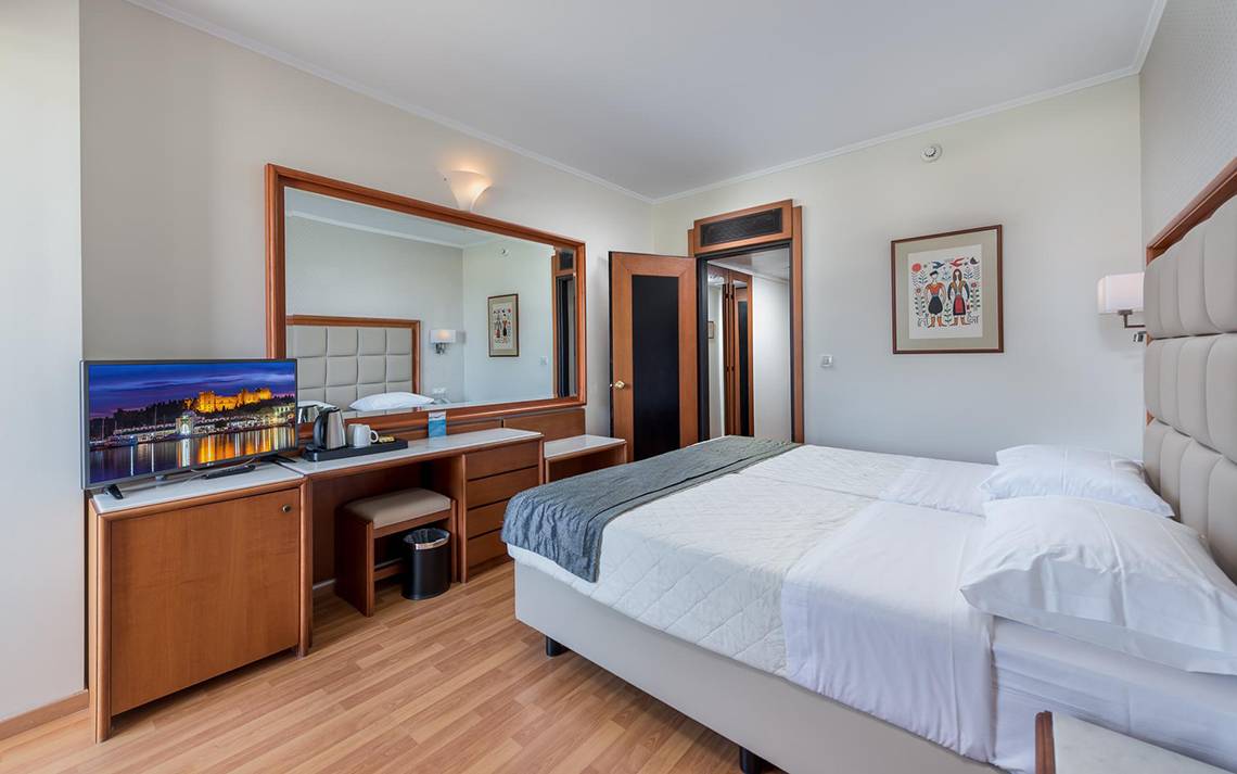 Innenansicht eines Hotelzimmers im Apollo Beach Hotel mit Fernseher, Schreibtisch und Bett.
