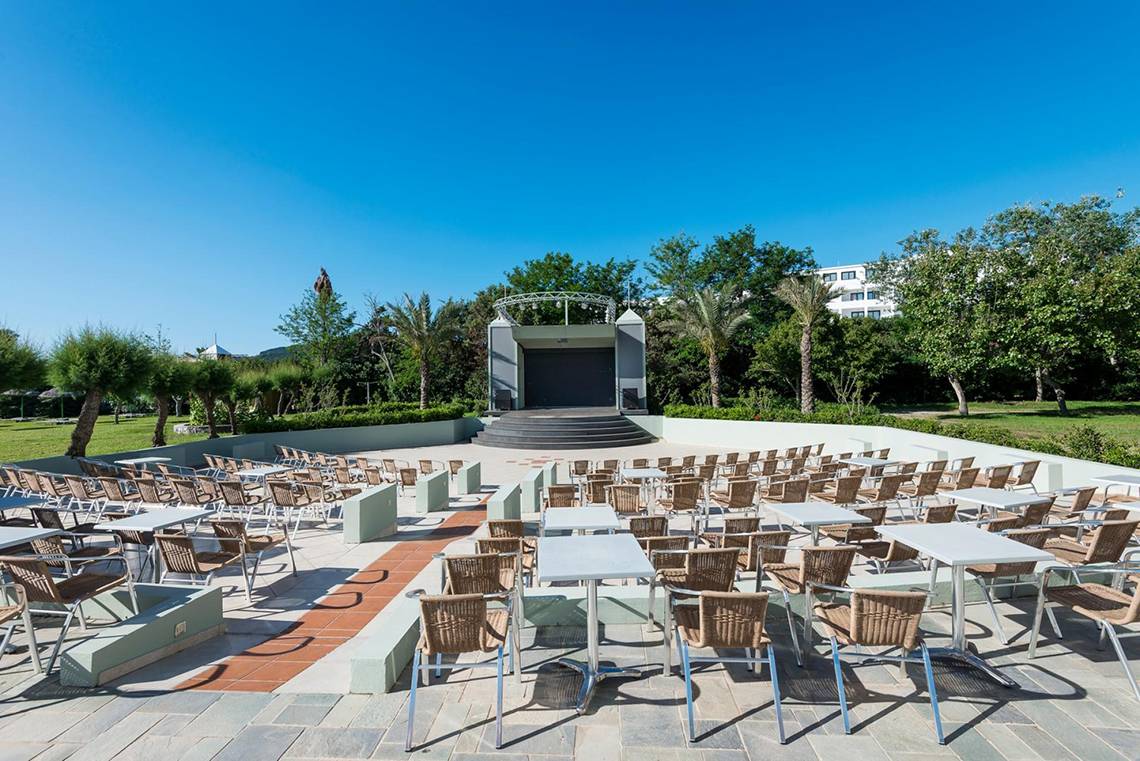 Apollo Beach Hotel mit Outdoor-Veranstaltungsort, vielen Tischen und Stühlen, Bühne und Bäumen im Hintergrund.