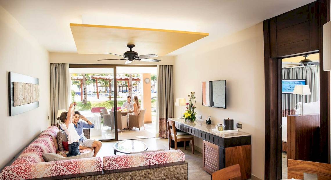 Innenansicht einer Suite im Barcelo Maya Grand Resort mit zwei Personen, die draußen sitzen und ein Mädchen, das die Hände hebt.
