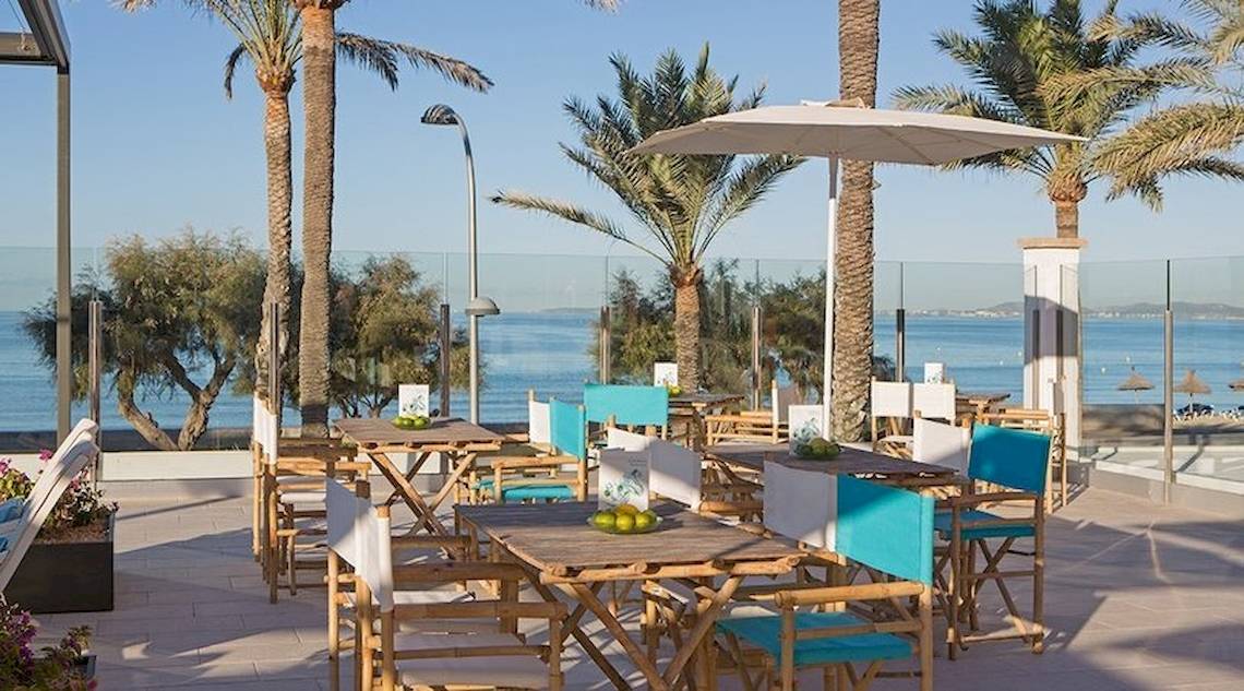 Außenterrasse des Universal Hotel Neptuno mit blauen Stühlen, Tischen und Sonnenschirmen mit Blick auf das Meer.