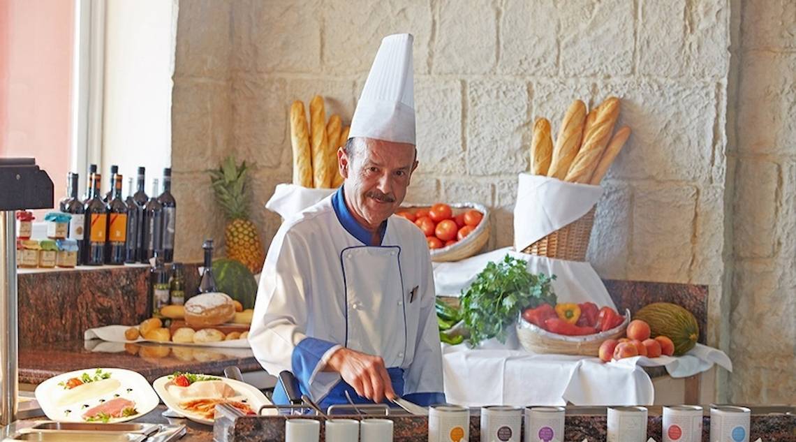 Koch in der Universal Hotel Neptuno Küche mit vorbereiteten Gerichten und Zutaten im Hintergrund.
