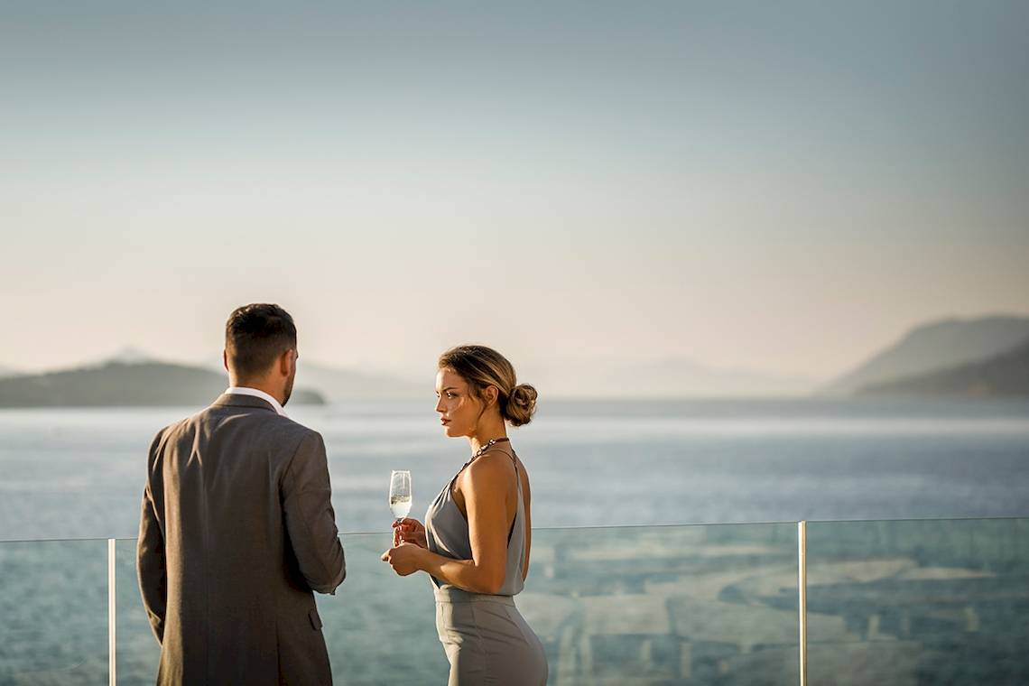 Mann und Frau auf einem Balkon mit Meerblick, der Mann mit einem Glas Wein und die Frau mit einem Glas Sekt.
