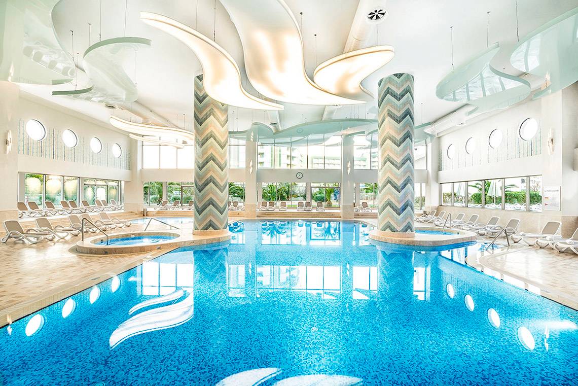 Ein großer Innenpool im Titanic Deluxe Lara Hotel mit Loungeliegen und modernen Leuchten.