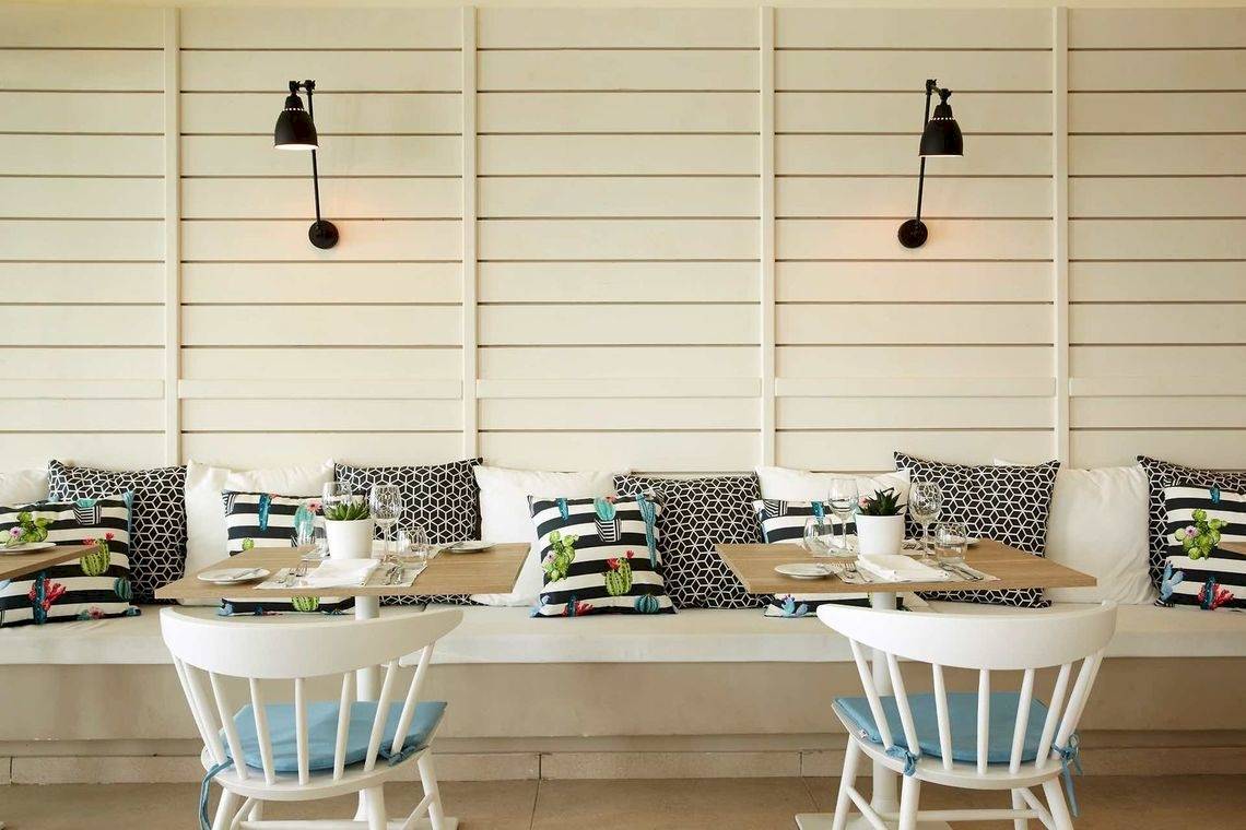 Innenansicht eines Restaurants im Pelekas Kloster mit weißen Bänken, Tischdecken und gemusterten Kissen.
