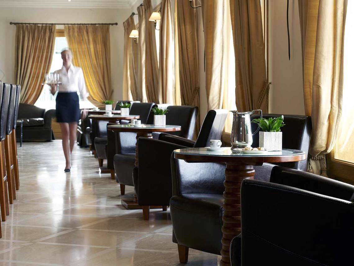 Innenansicht des Restaurants des Mitsis Rinela Beach Hotels mit einer Kellnerin, die eine Tablett mit Gläsern trägt.