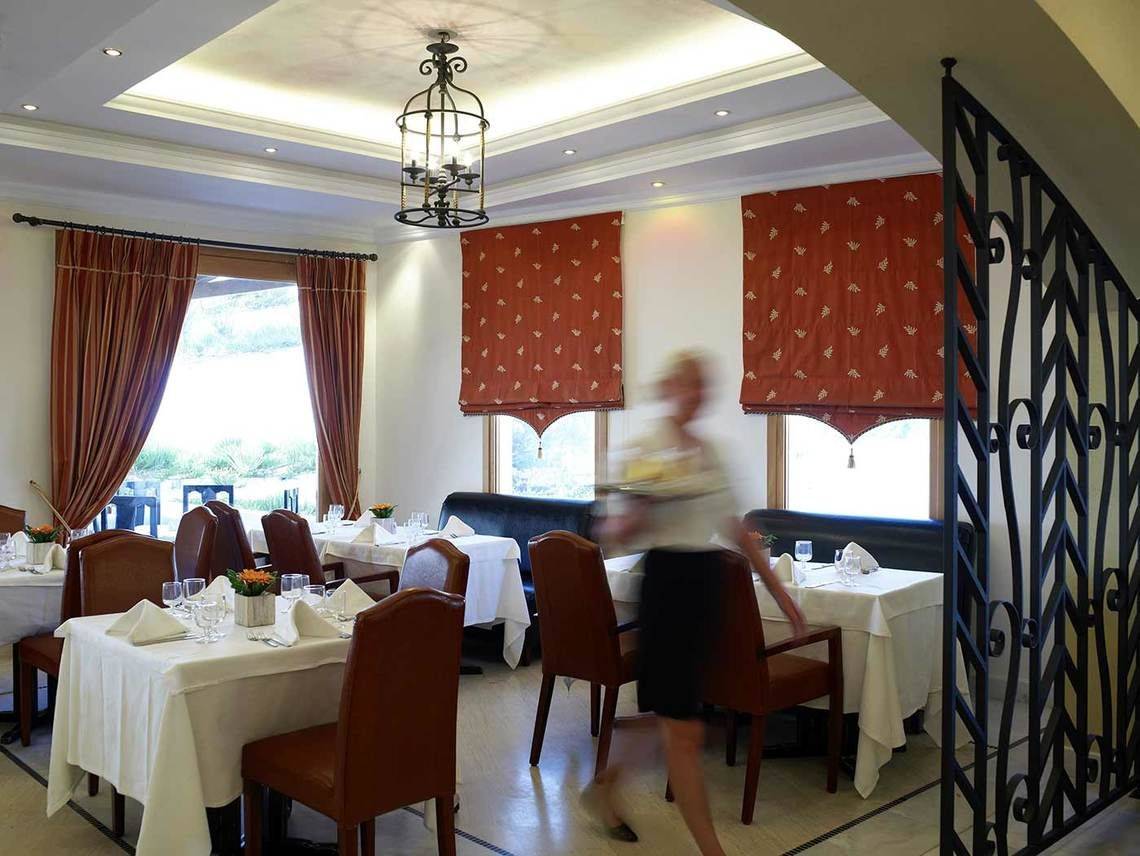 Innenansicht des Restaurants des Mitsis Rinela Beach Hotels mit Tisch und Stühlen.