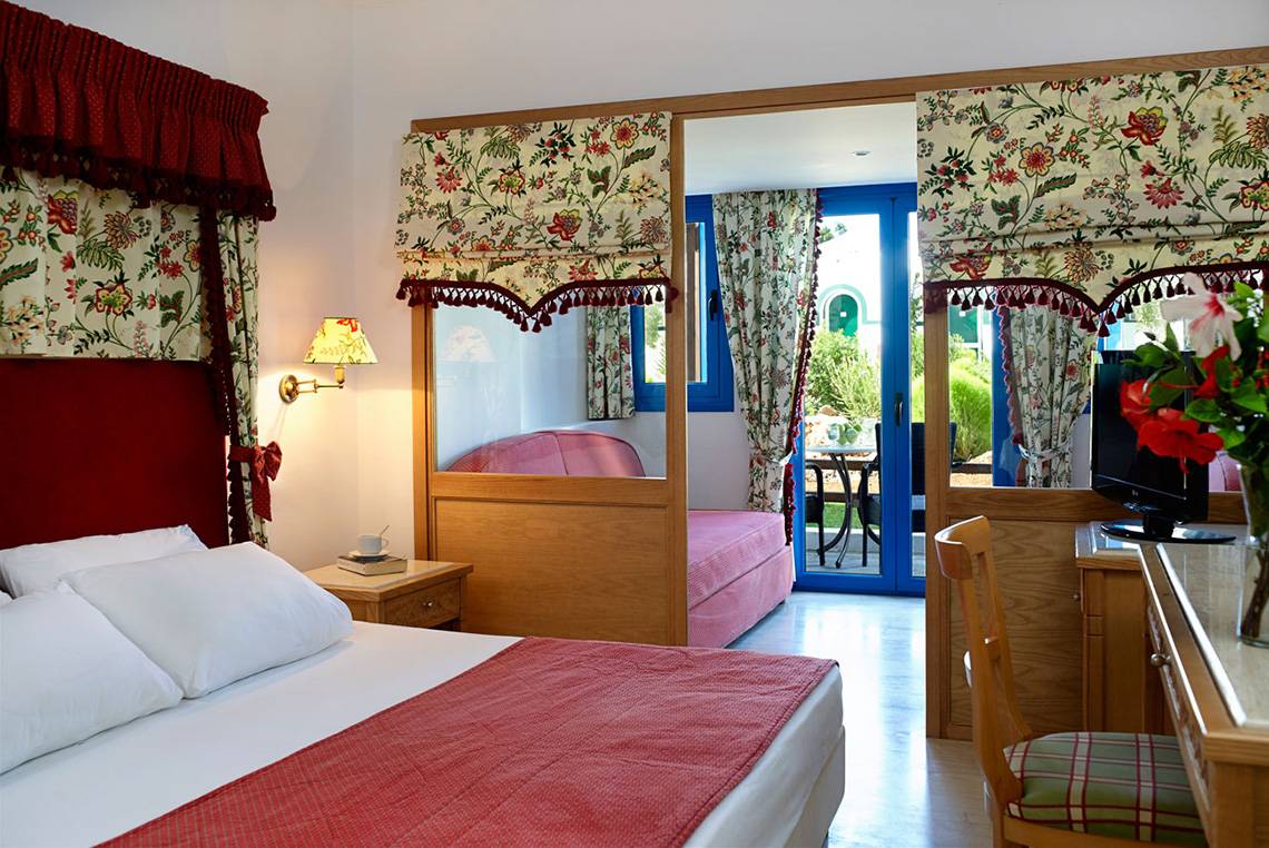 Ein Schlafzimmer im Mitsis Rinela Beach Hotel mit Doppelbett, Nachttisch, Sessel und Balkon.