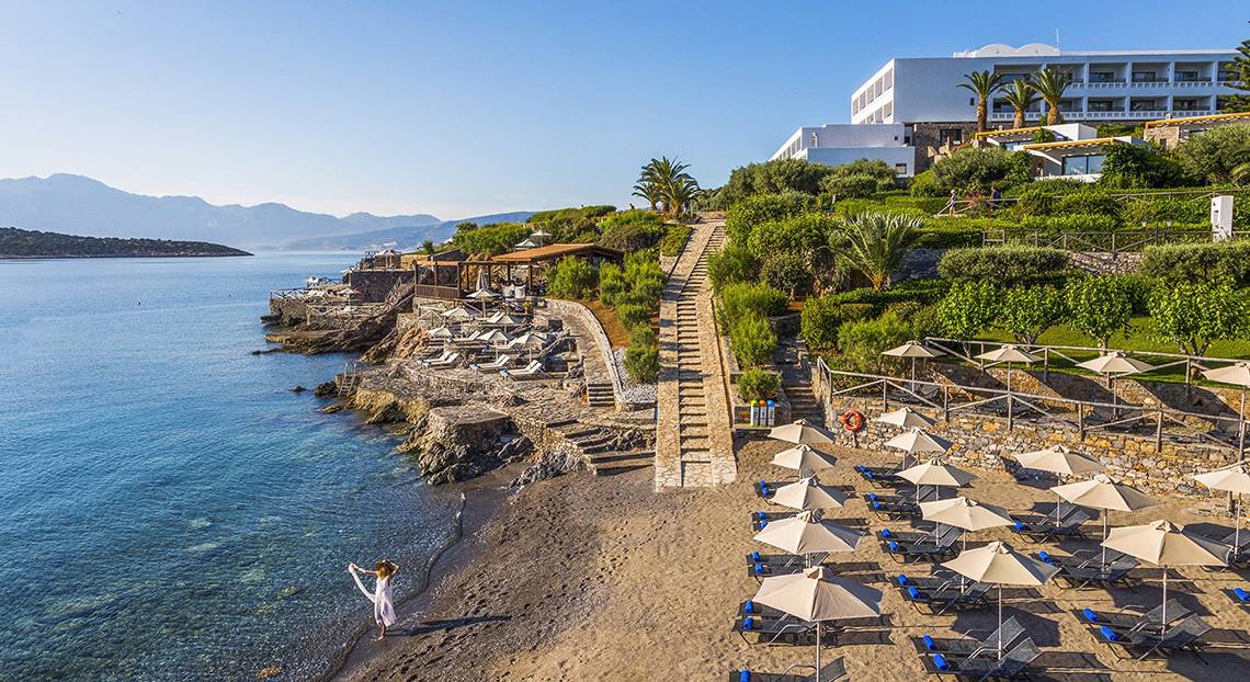 Luftaufnahme des Minos Palace Hotel & Suites mit Blick auf den Strand, Liegestühlen und Sonnenschirmen.