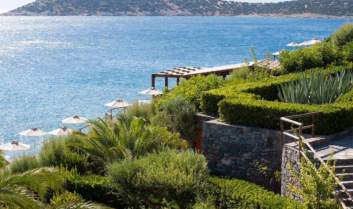 Außenbereich des Minos Palace Hotel & Suites mit Blick auf das Meer und gesäumt von Palmen und Sträuchern.