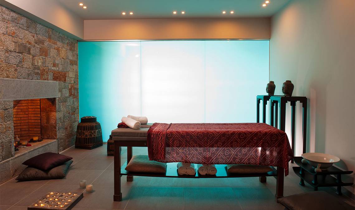 Spa-Bereich im Minos Palace Hotel & Suites mit Massagetisch, Kissen, Kamin und Glaswand.