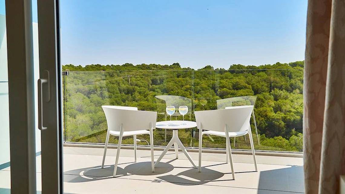 Blick von einem Hotelbalkon auf einen Tisch mit zwei Weingläsern und zwei weißen Stühlen mit Blick auf einen Wald.