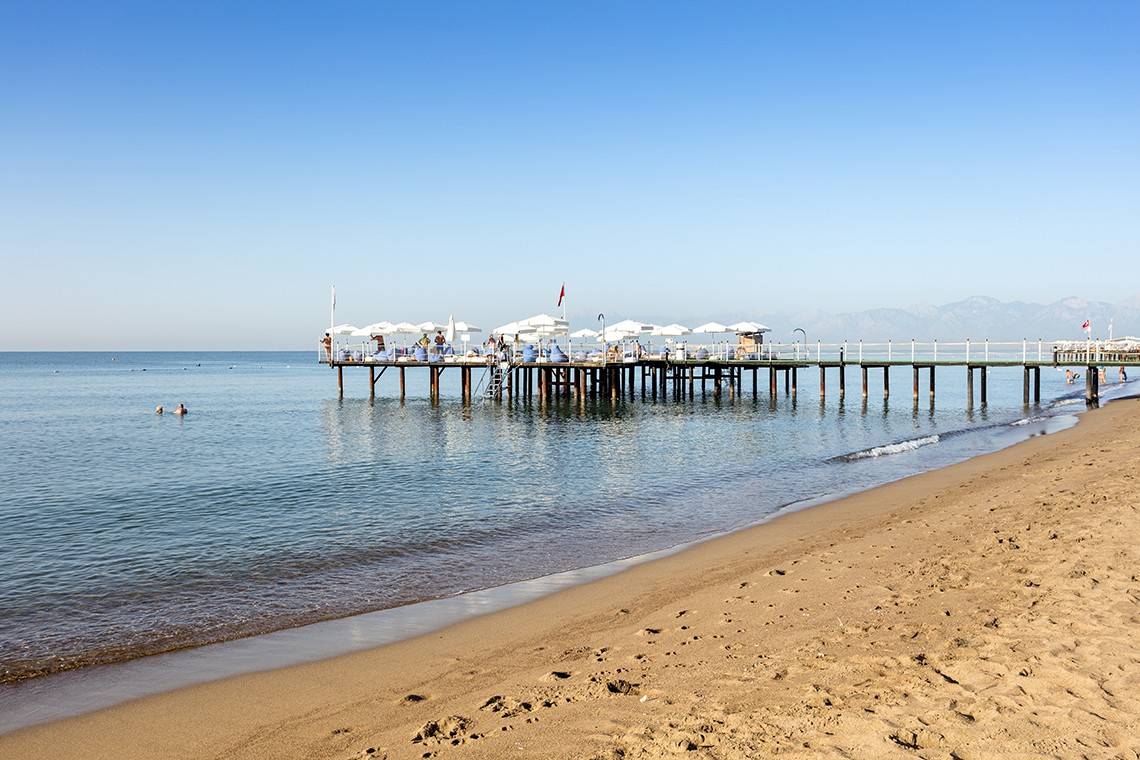 Eine Strandszene mit einem Pier im Hintergrund, wo einige Personen unter weißen Sonnenschirmen sitzen.