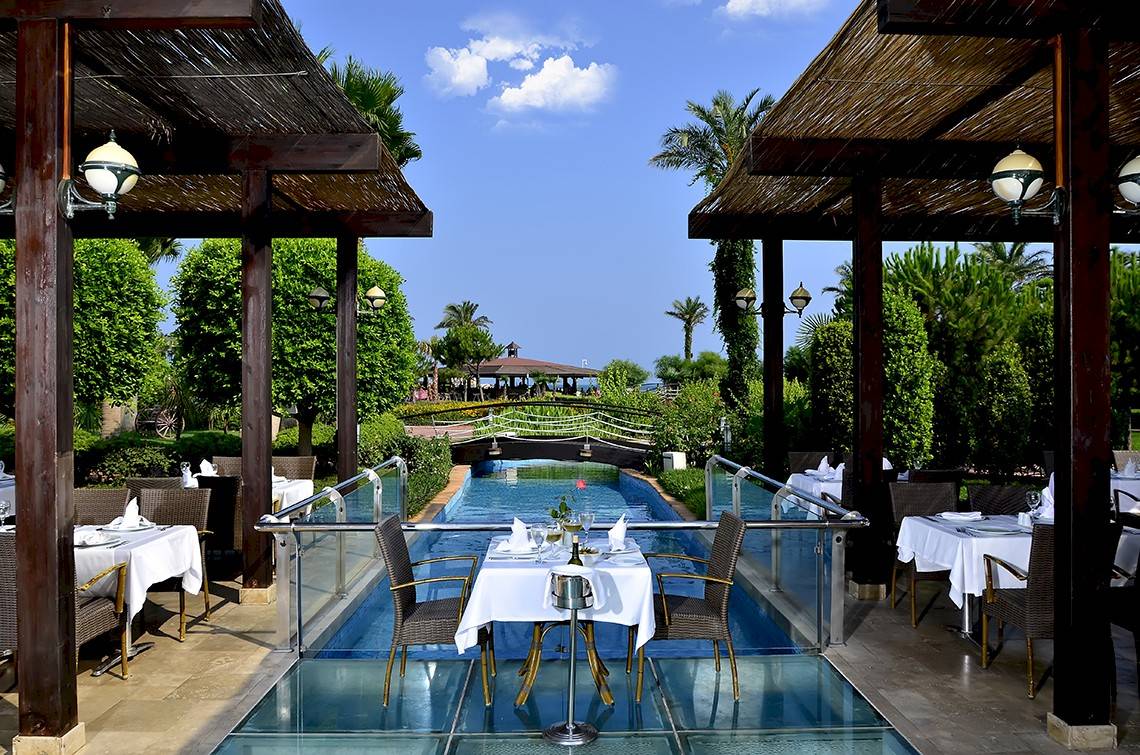 Pool-Terrasse im Miracle Resort mit Tisch und Stühlen, umgeben von Pflanzen und einer Brücke.