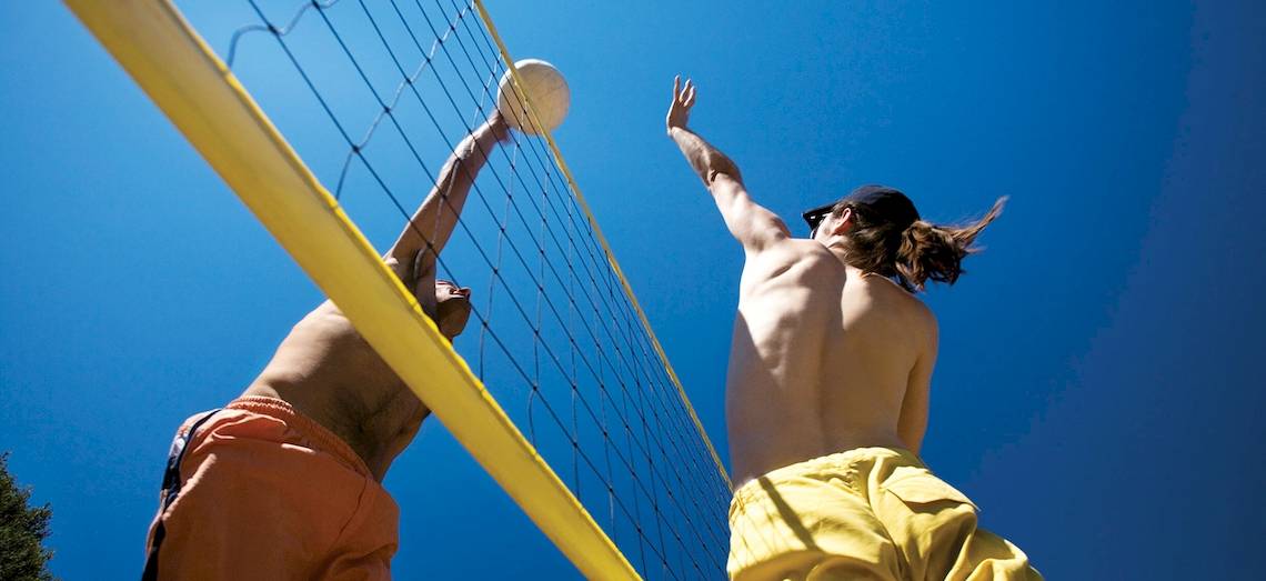 Zwei Männer spielen Beachvolleyball mit gelben Netz und blauem Himmel im Hintergrund.