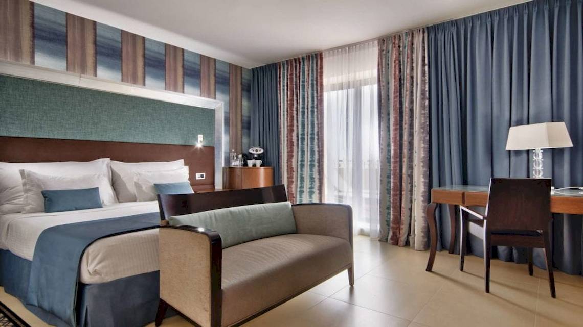 AX The Palace Hotel Zimmer mit Bett, Couch, Schreibtisch und Lampe