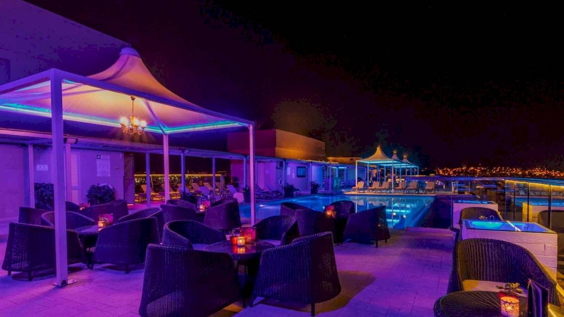 Die Pool-Lounge-Area des AX The Palace Hotels bei Nacht mit lila Beleuchtung und runden Tischen.
