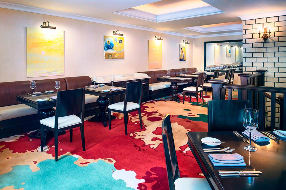 Restaurant im Sheraton Jumeirah Beach Resort mit gemustertem Teppich, dunklen Tischen und Stühlen.