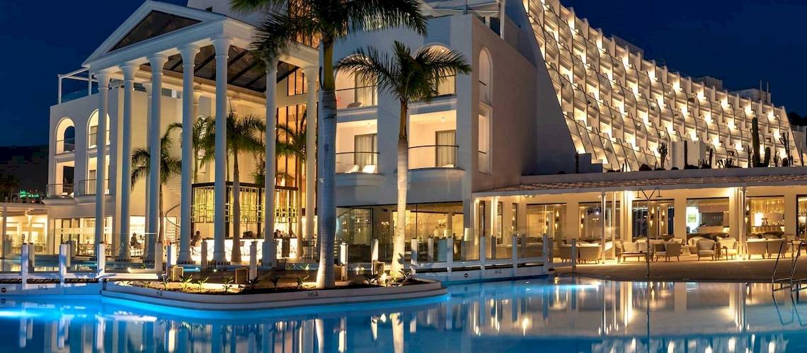 Die Guayarmina Princess Hotel Fassade ist beleuchtet und zeigt eine elegante Architektur mit Palmen und einem Pool vor dem Gebäude.
