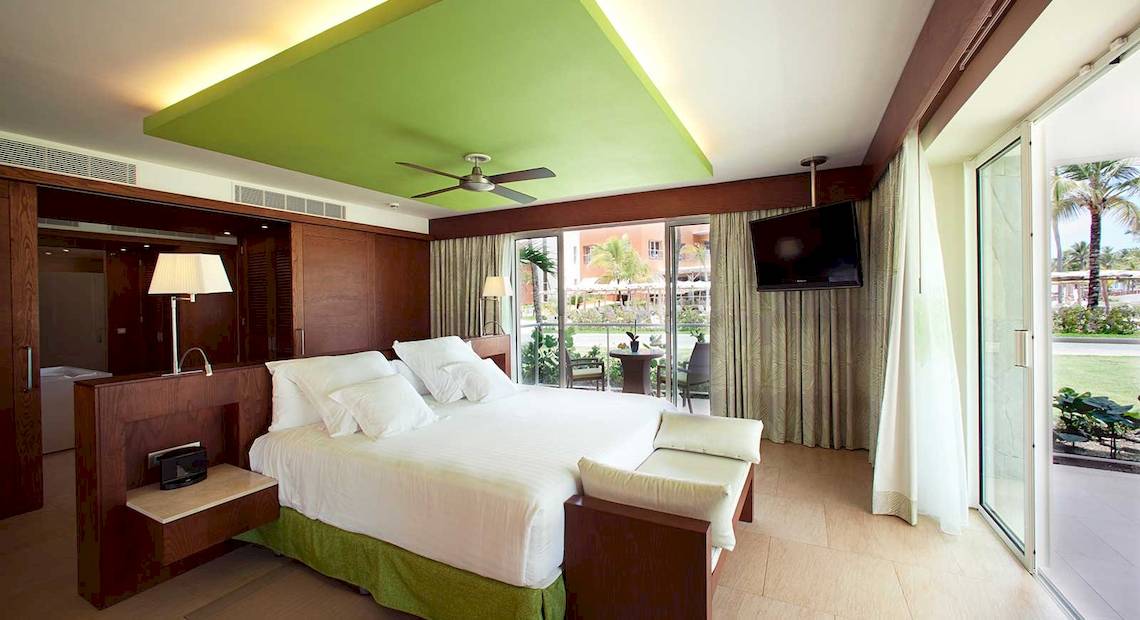 Hotelzimmer mit großem Bett, grüner Decke, Fernseher, Glastüren und Blick auf den Garten.