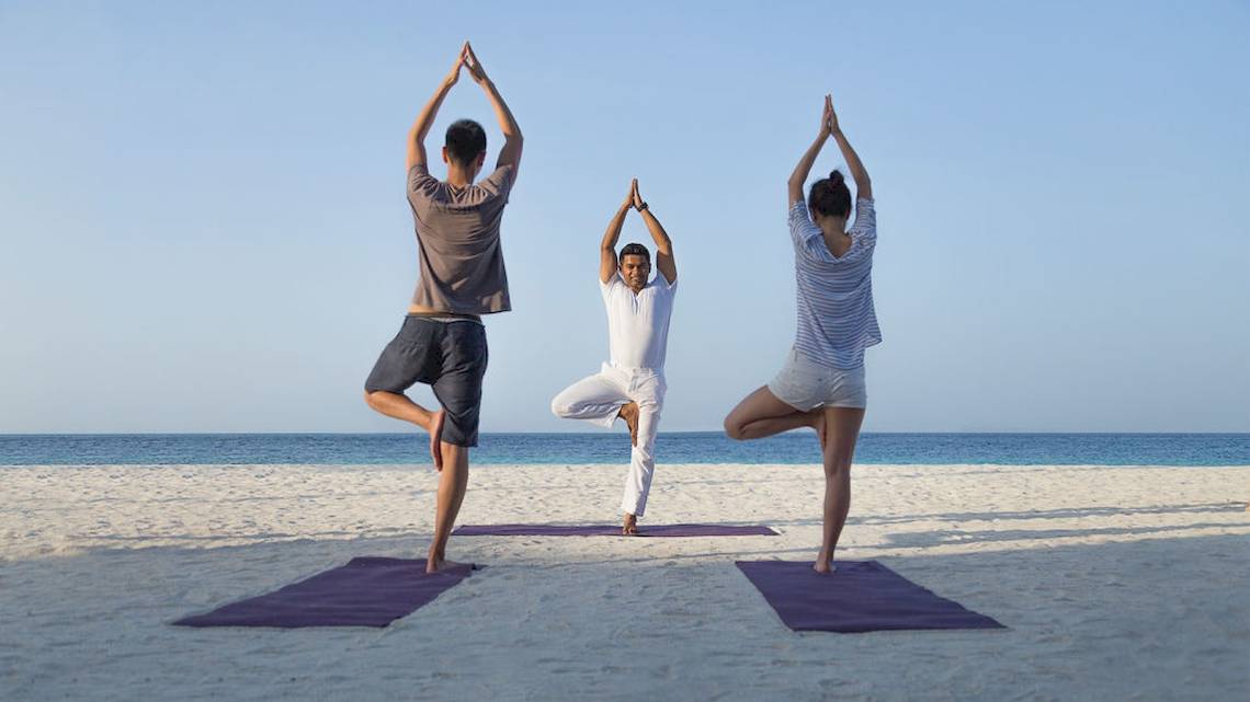 Drei Personen machen Yoga auf dem Strand des Komandoo Island Resorts & Spa.