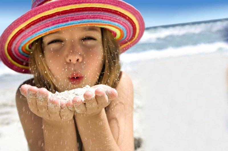 Kleines Mädchen am Strand mit Regenbogenhut, das Sand durch die Hände rieseln lässt.