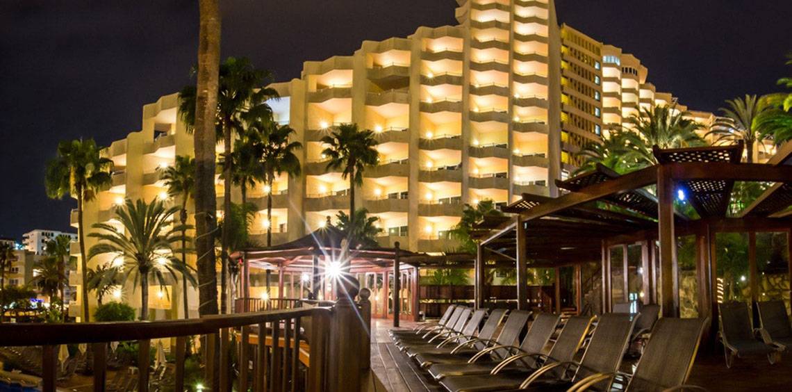 Corallium Dunamar Garden Hotel bei Nacht mit vielen leuchtenden Fenstern und Lounge-Bereich davor.