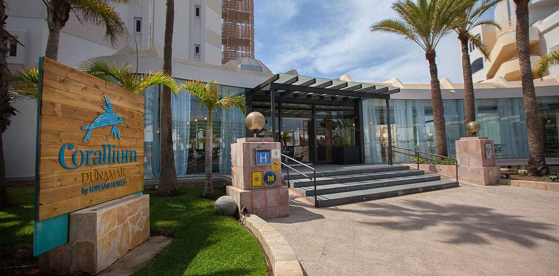 Eingang des Corallium Dunamar Garden Hotels mit Holzschild und Palmen