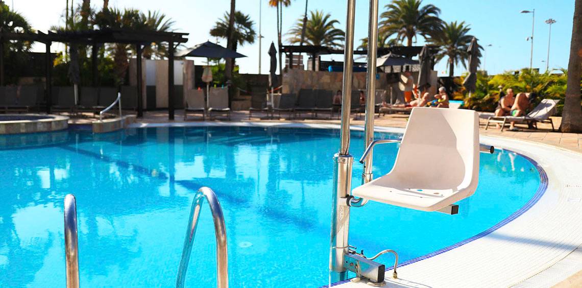 Ein behindertengerechter Stuhl steht am Rande des Schwimmbeckens im Corallium Dunamar Garden by Lopesan Hotels.