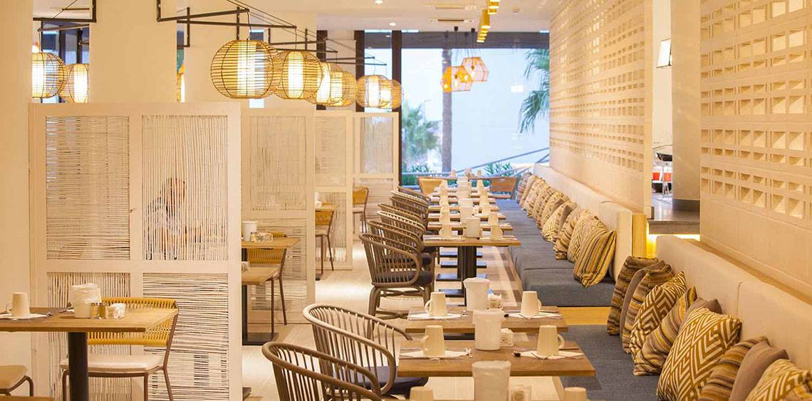 Innenansicht des Restaurants des Corallium Dunamar Garden by Lopesan Hotels mit Tisch, Stühlen, Lampen und Wandgestaltung.