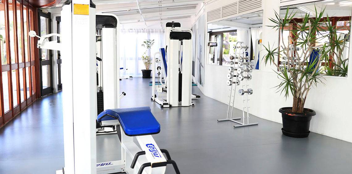 Innenansicht des Fitnesscenters im Corallium Dunamar Garden by Lopesan Hotels mit Geräten und Pflanzen.